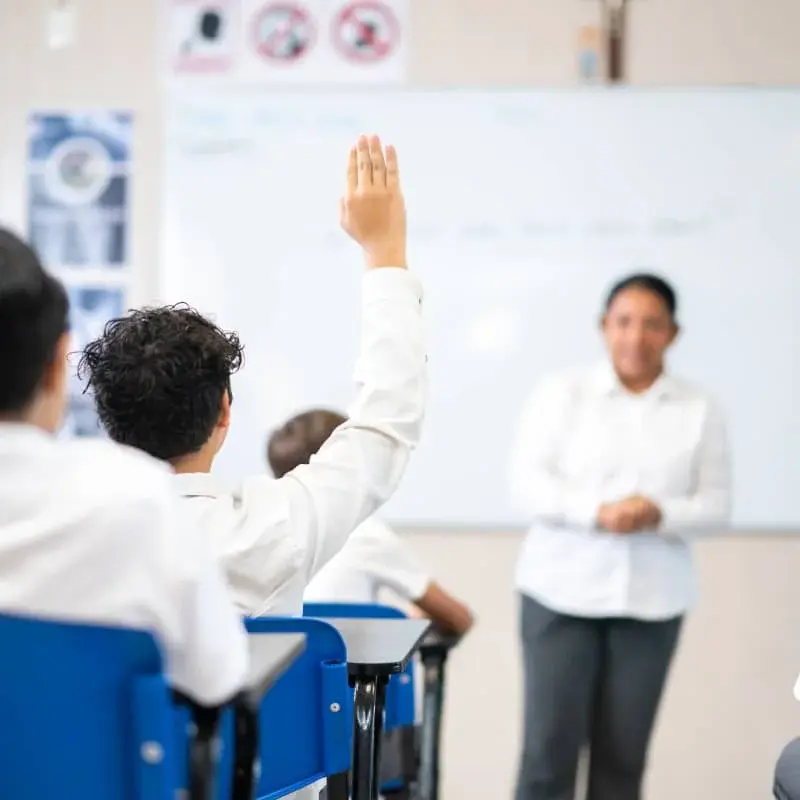 Estudiante levanta la mano en clase mientras el docente enseña frente a la pizarra blanca